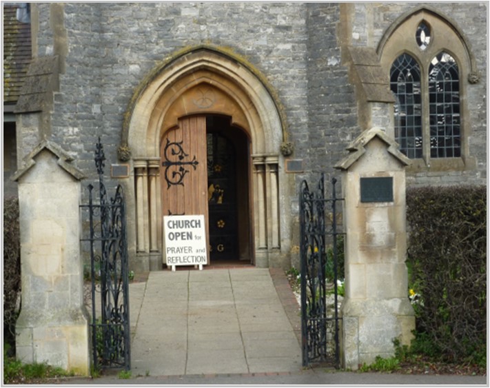 Open iron gates in front of open church door with sign saying Church Open for prayer and reflection. 